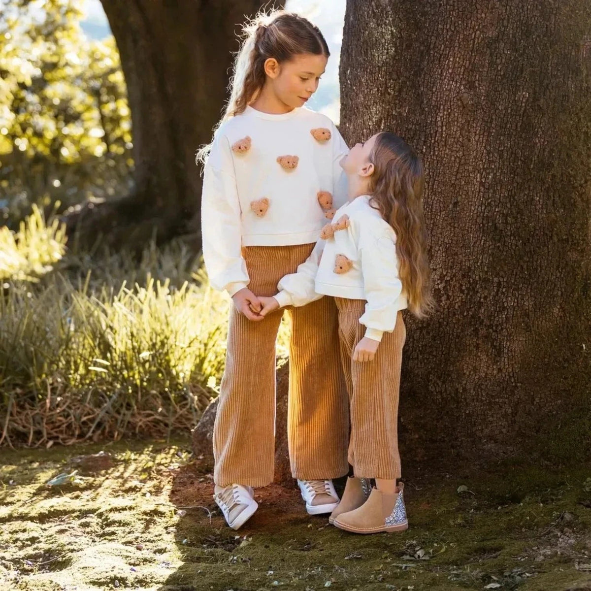 Twee meisjes dragen Pierrot Outlet off-white sweaters met pluizige teddybeer-applicaties en bruine ribfluwelen broeken.