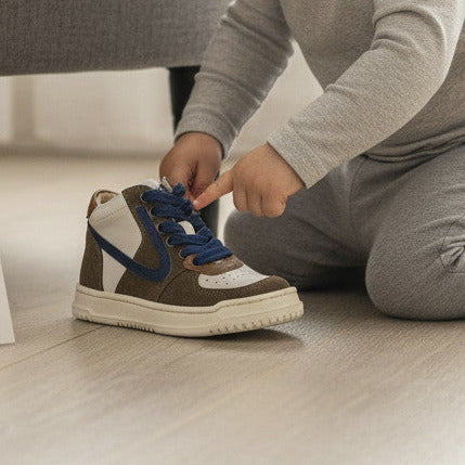 Peuter speelt met Pierrot Outlet hoge sneakers in bruin, wit en blauw, gemaakt van suède en leer op een houten vloer.