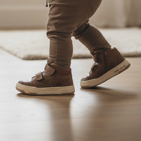 Peuter draagt cognacbruine Pierrot Outlet leren sneakers met klittenband en ster-accent in een lichte kinderkamer.