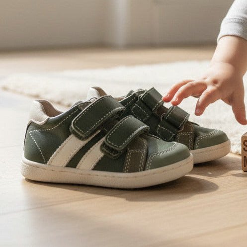 Pierrot Outlet olijfgroene leren kindersneakers met klittenband en witte accenten, zittend op een houten vloer.